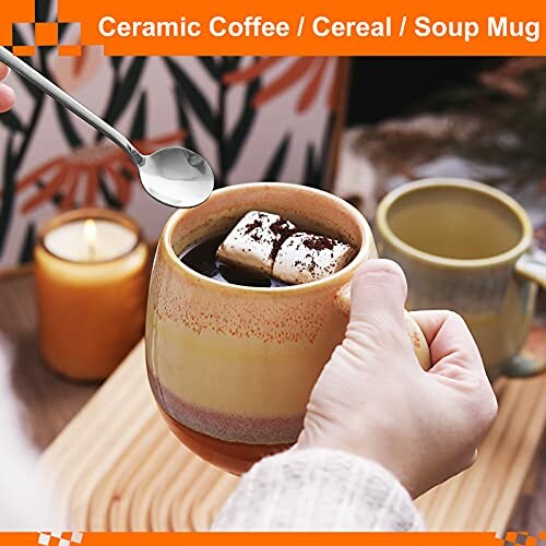 Hand holding a ceramic coffee mug with marshmallows and a spoon.